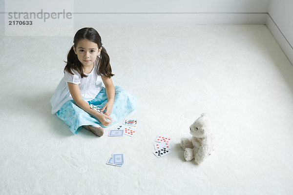 Mädchen sitzt auf dem Boden und spielt Kartenspiel mit Teddybär  schaut in die Kamera.