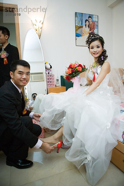 Bräutigam setzt Schuh auf Brautfuß im Schlafzimmer  beide lächelnd vor der Kamera  volle Länge