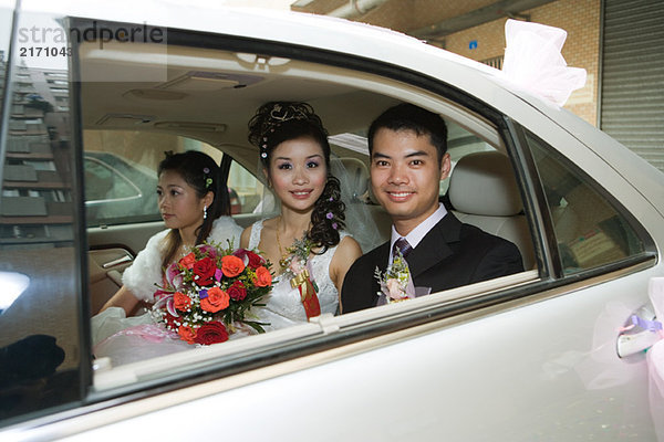 Braut und Bräutigam im Auto mit Brautjungfer  lächelnd vor der Kamera