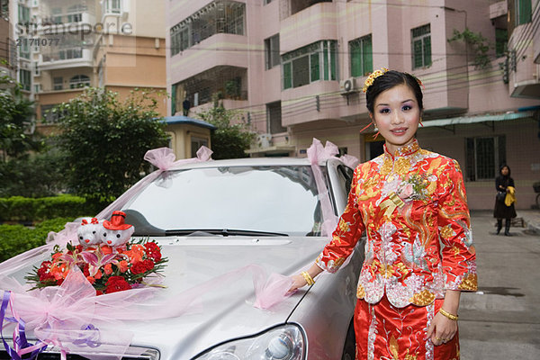 Braut in traditioneller chinesischer Kleidung  neben dem geschmückten Auto stehend  lächelnd vor der Kamera.