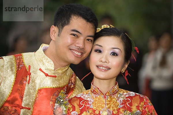 Braut und Bräutigam lächeln gemeinsam vor der Kamera  beide in traditioneller chinesischer Kleidung.