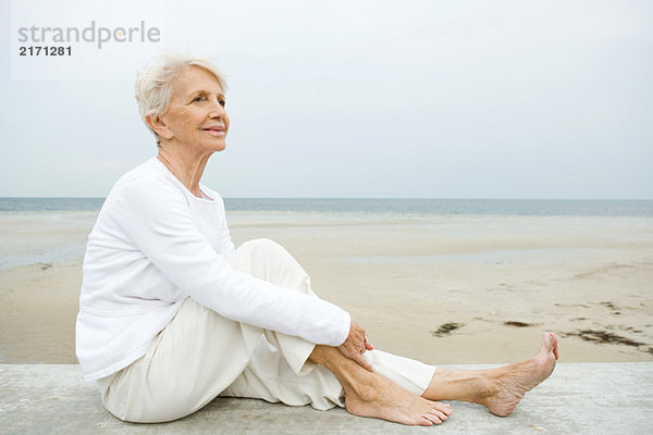 Seniorin auf niedriger Wand sitzend  Strand im Hintergrund  volle Länge
