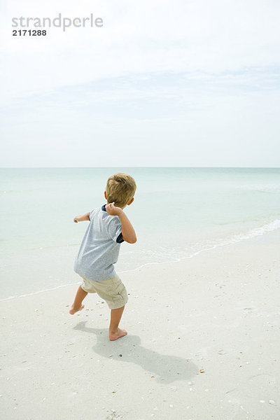 Junge am Strand wirft unsichtbares Objekt zum Meer  Rückansicht  volle Länge
