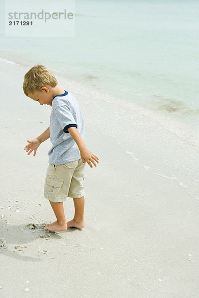 Kleiner Junge  der am Strand steht und Zehen in Sand gräbt.