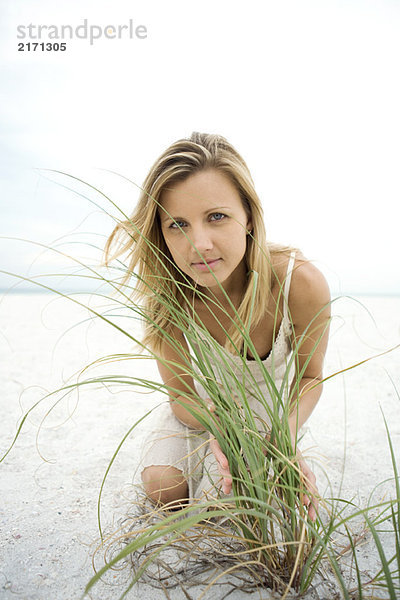 Frau kniend am Strand  durch hohes Gras blickend