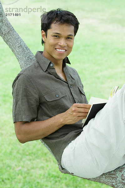 Mann ruht auf Baumzweig  hält Buch  lächelt in die Kamera