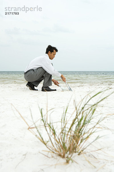 Mann hockt am Strand  fegt Sand in die Kehrschaufel  Seitenansicht