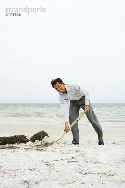 Mann am Strand schaufelt Sand mit Schaufel  lächelt in die Kamera  volle Länge