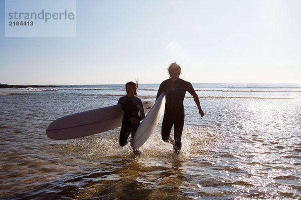 Ein Paar  das lächelnd durchs Wasser läuft und Surfbretter trägt.