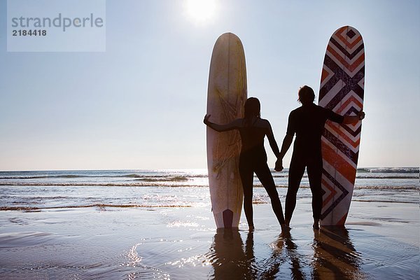 Paar am Strand stehend mit Surfbrettern an den Händen.