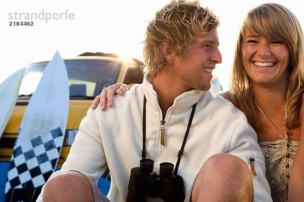 Ein Paar sitzt am Strand und lächelt mit Van und Surfbrettern im Hintergrund.