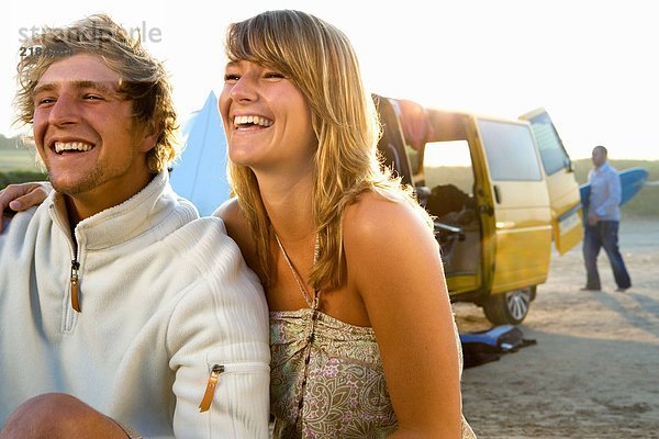Ein Paar sitzt am Strand und lächelt mit einem Mann mit Surfbrett im Hintergrund.
