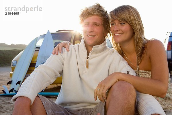 Ein Paar sitzt am Strand und lächelt mit Van und Surfbrettern im Hintergrund.