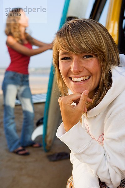 Frau am Strand lächelnd mit einer anderen Frau im Hintergrund  die das Surfbrett hält.