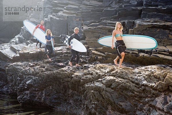 Vier Leute tragen Surfbretter auf großen Felsen.