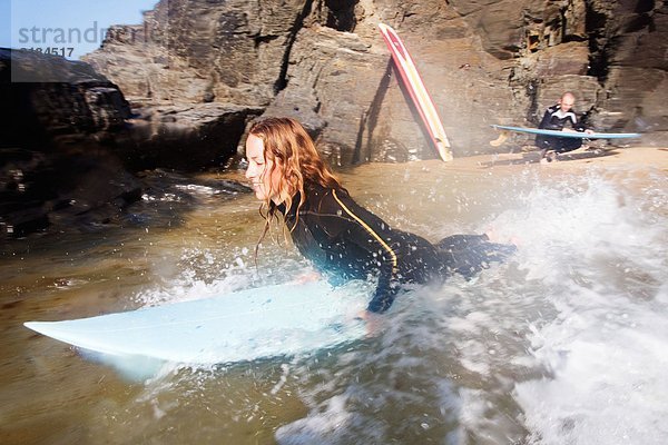 Frau auf Surfbrett lächelnd mit Mann im Hintergrund mit Surfbrett.