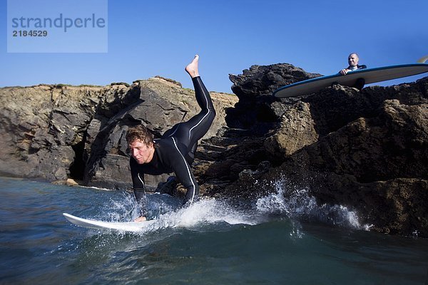 Ein Mann springt auf das Surfbrett  während ein anderer Mann auf Steinen hinter ihm sitzt.