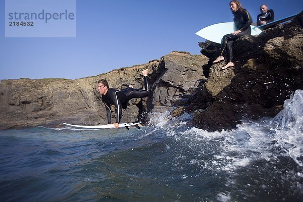 Mann  der auf dem Surfbrett ins Wasser springt und zwei Leute auf großen Felsen lächelnd sitzen.