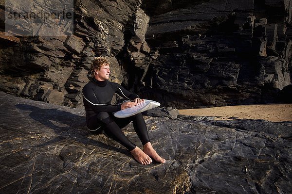 Mann sitzt auf großem Felsen im Neoprenanzug mit Surfbrett auf dem Schoß.