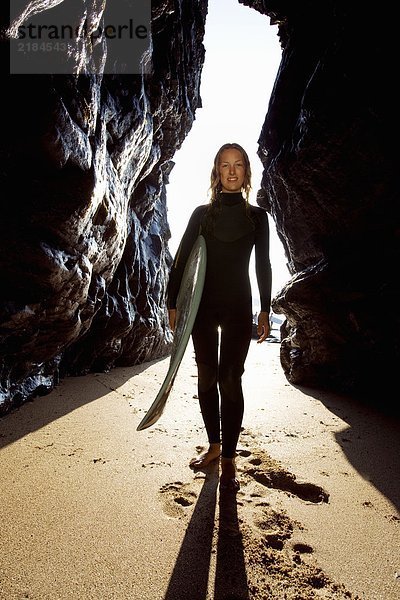 Frau steht mit Surfbrett zwischen großen Felsen und lächelt.