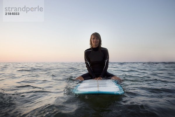 Frau sitzt auf dem Surfbrett im Wasser und lächelt.