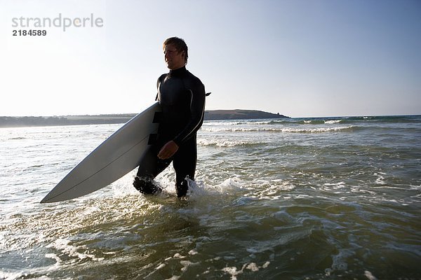 Mann geht mit einem Surfbrett im Wasser und lächelt.