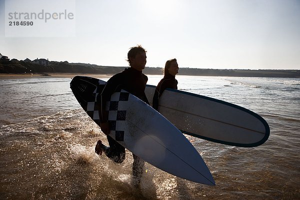 Ein Paar  das mit lächelnden Surfbrettern durchs Wasser läuft.