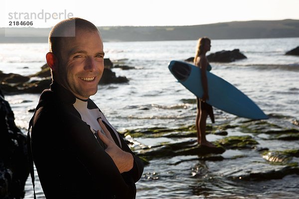 Mann steht lächelnd mit einer Frau  die das Surfbrett im Hintergrund hält.