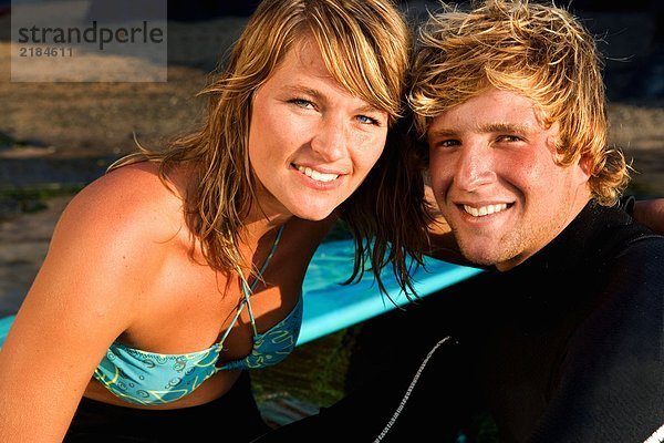 Paar sitzend zusammen lächelnd mit Surfbrett im Hintergrund.
