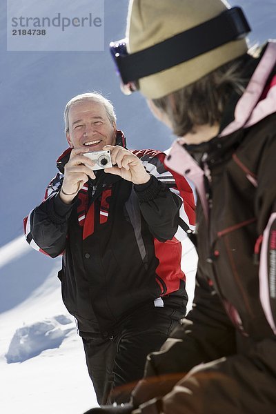 Reifer Mann in Skibekleidung fotografiert Frau am Berg