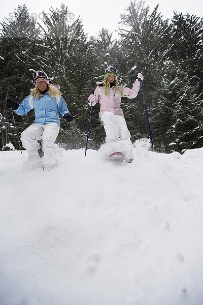 Tiefblick auf zwei junge blonde Frauen  die mit Schneeschuhen den kleinen Hügel im Wald hinuntergehen.