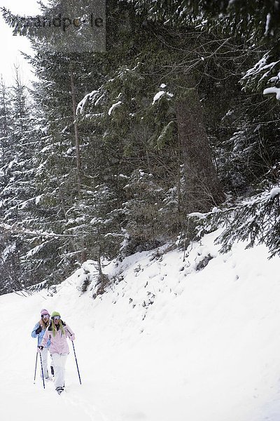 Zwei junge blonde Frauen  die mit Schneeschuhen den kleinen Hügel im Wald hinuntergehen.