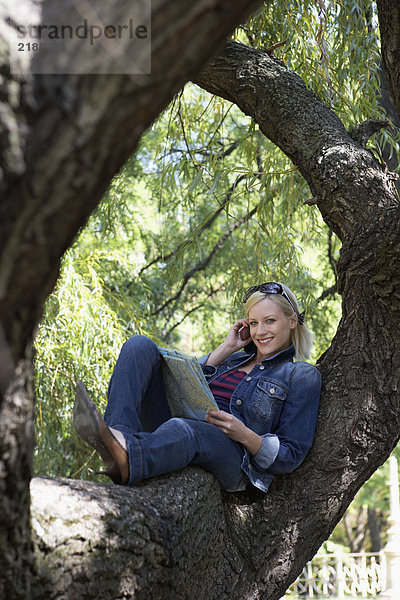 Frau sitzt auf einem Baum auf dem Handy und lächelt.