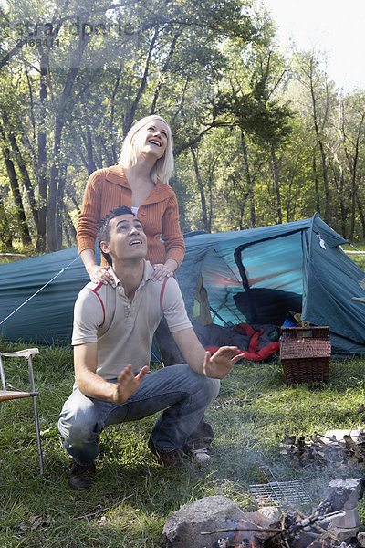 Ein Paar auf dem Campingplatz schaut lächelnd auf den Rauch der Feuerstelle.