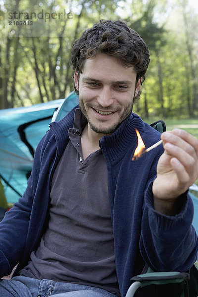 Ein Mann  der auf dem Campingplatz sitzt und lächelnd ein Streichholz hält.