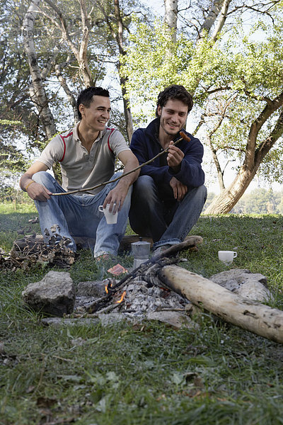 Zwei Männer kochen Hot Dogs über einer Feuerstelle und lächeln.