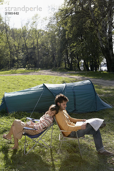 Ein Paar sitzt Rücken an Rücken auf dem Campingplatz mit dem Mann  der eine Zeitung liest.