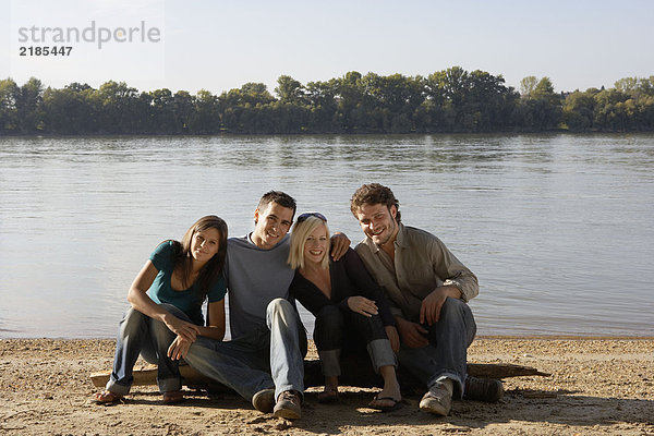 Vier Freunde sitzen auf einem Baumstamm am See und lächeln.