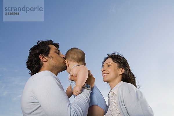 Ein Paar hält ein Baby draußen  während der Mann es küsst.