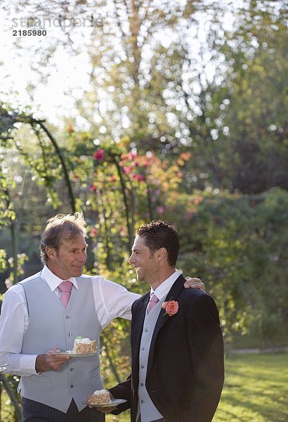 Reife Vater und Bräutigam halten Teller von Hochzeitstorte im Garten