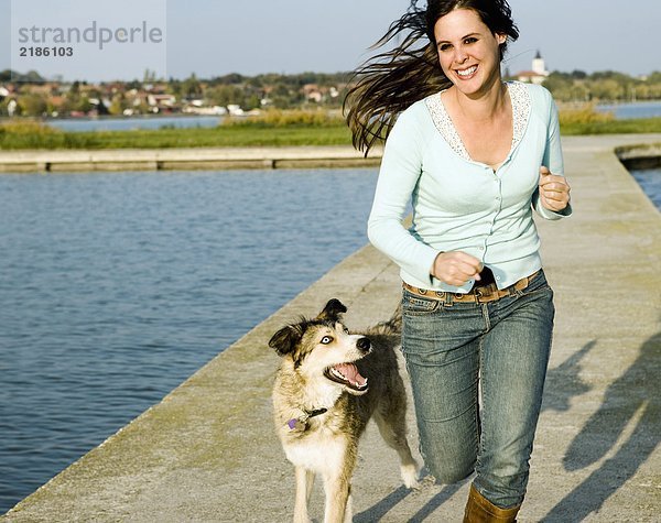 Frau läuft mit Hund an einem See entlang.