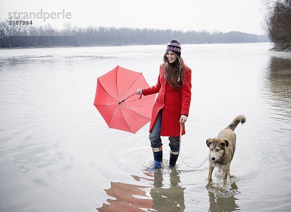 Junge Frau hält Regenschirm  watet im Fluss mit Hund  lächelt