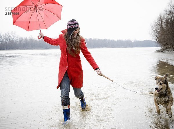 Junge Frau hält Regenschirm  läuft mit Hund im Fluss  lächelt