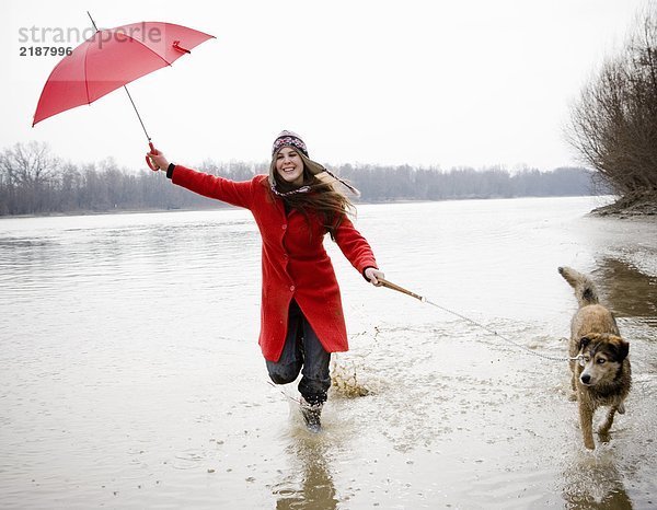 Junge Frau hält Regenschirm  läuft mit Hund im Fluss  lächelt