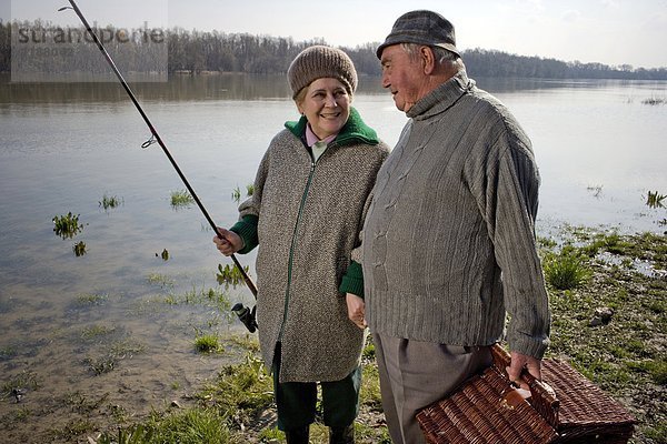 Seniorenpaar mit Picknickkorb und Angelrute am Fluss  lächelnd