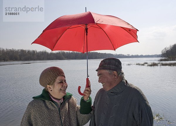 Seniorenpaar steht am Fluss und teilt sich den Regenschirm  lächelnd