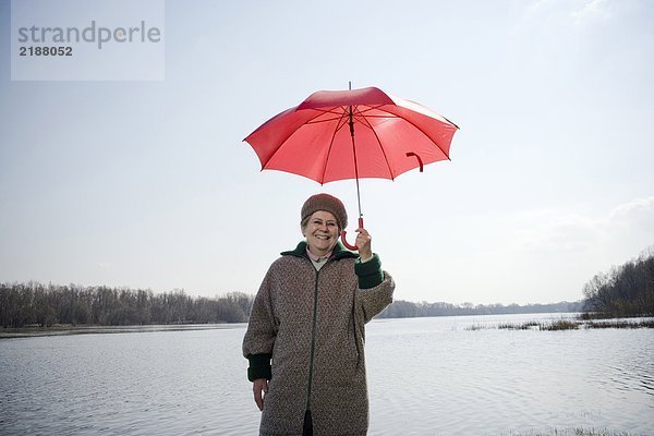 Seniorin am Fluss stehend mit rotem Regenschirm  lächelnd  Portrait