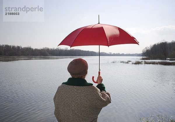 Seniorin am Fluss stehend mit rotem Regenschirm  Rückansicht