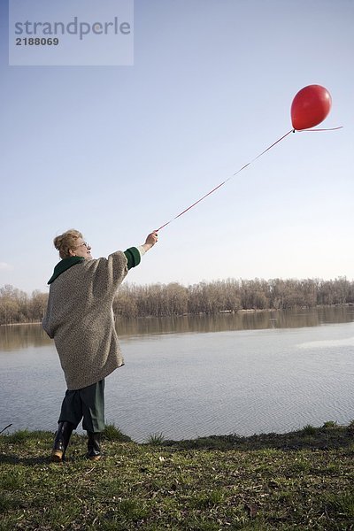 Seniorin am Fluss stehend mit rotem Ballon  Rückansicht