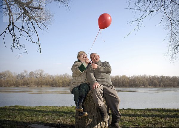 Seniorenpaar am Fluss sitzend mit rotem Ballon  lächelnd